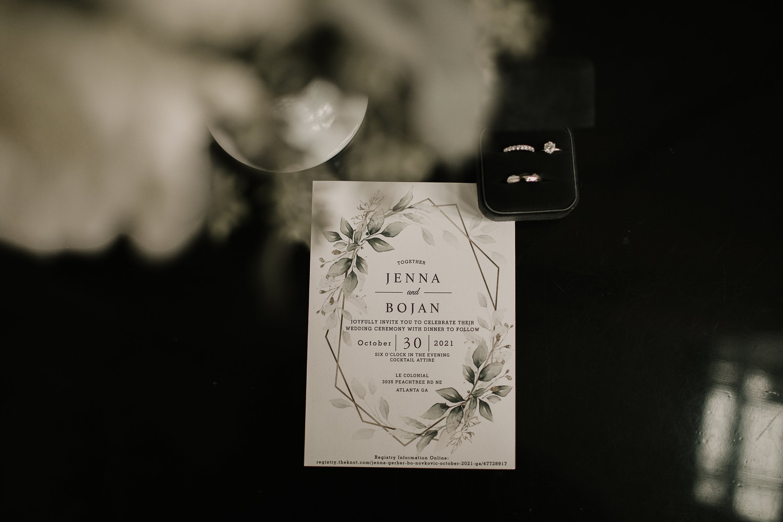 wedding detail flat lay image of ring, flowers and engagement invite on a black shadowed background.